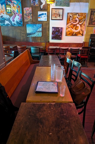 A Toronto bar bench, table and chairs with various artworks in the background. A pitcher, glasses and a menu are on the center table.
